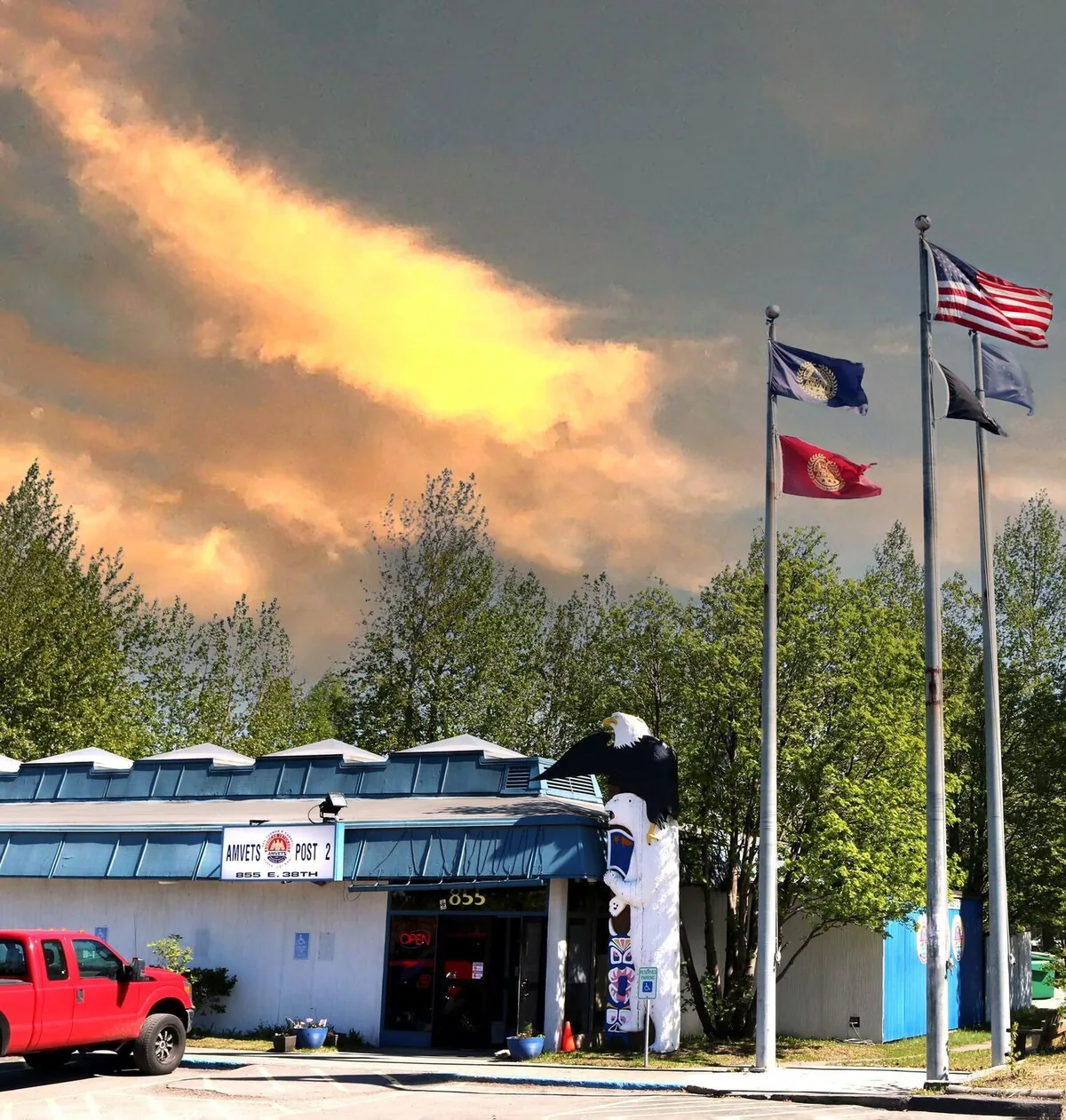 Front of the AMVETS Post 2 building in Anchorage beneath American and service flags, with an eagle and totem sculpture at the entrance